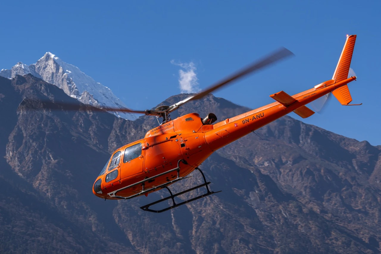 Bright orange Annapurna Helicopters Airbus H125 with registration 9N-ANU flying against a backdrop of steep rocky hills and snow-capped Himalayan peaks in Nepal.