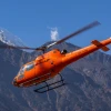 Bright orange Annapurna Helicopters Airbus H125 with registration 9N-ANU flying against a backdrop of steep rocky hills and snow-capped Himalayan peaks in Nepal.